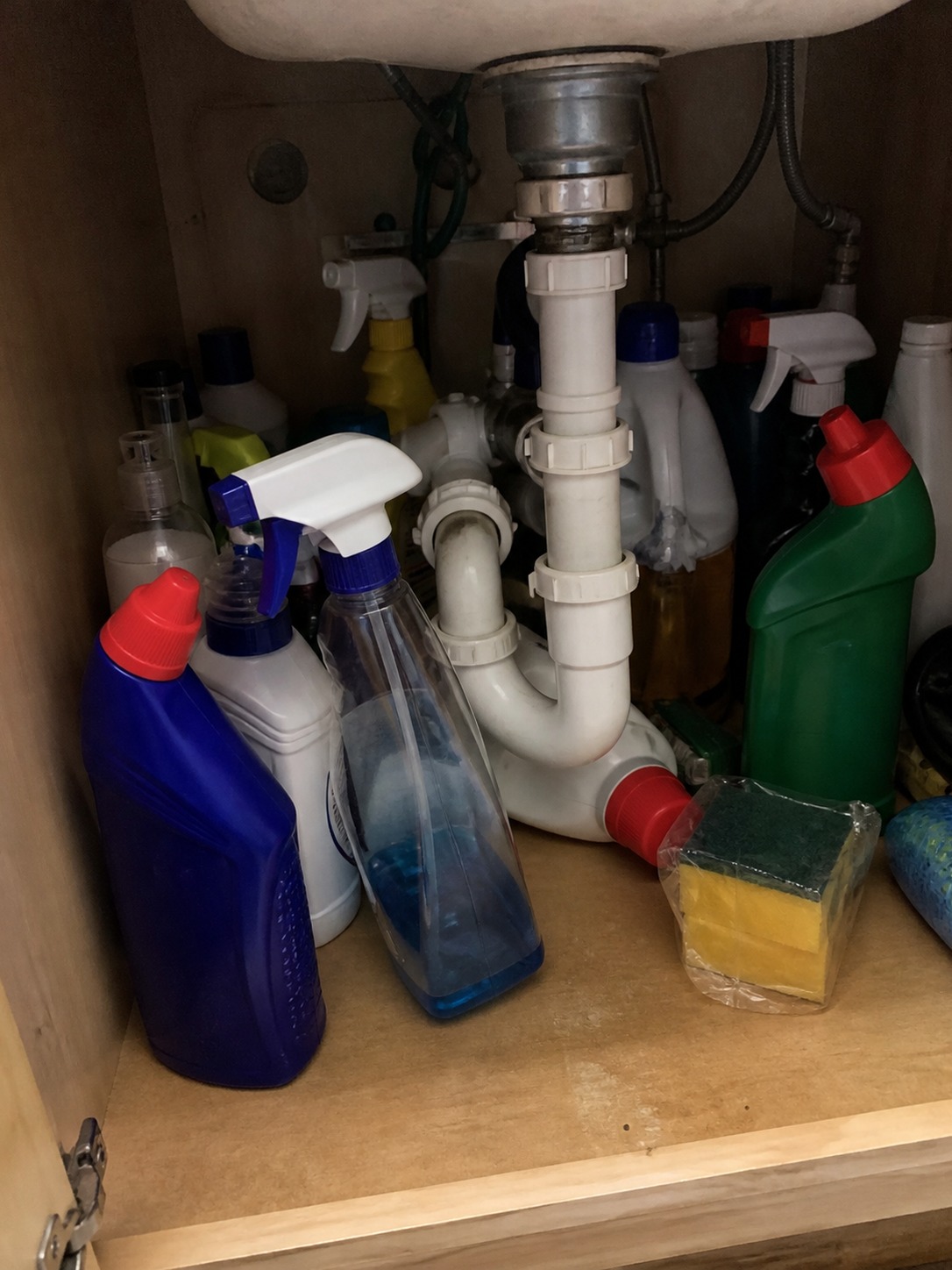 Cluttered under-sink cabinet with cleaning bottles crowded around plumbing pipes